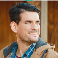 Smiling man with dark hair wearing a jacket and plaid shirt.