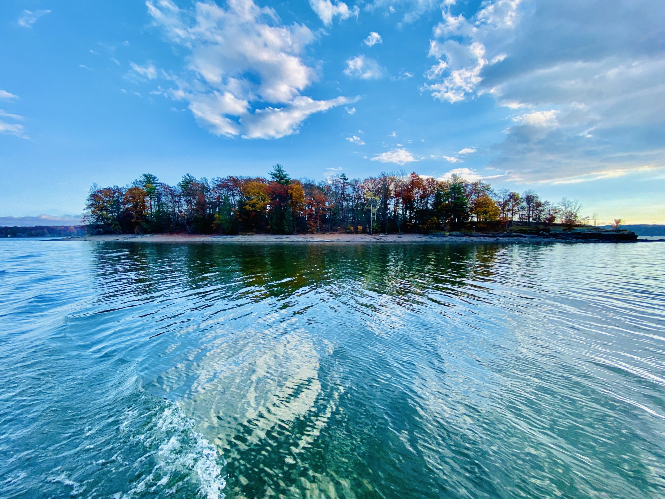 Calm lake with a colorful tree-lined shore under a blue sky.