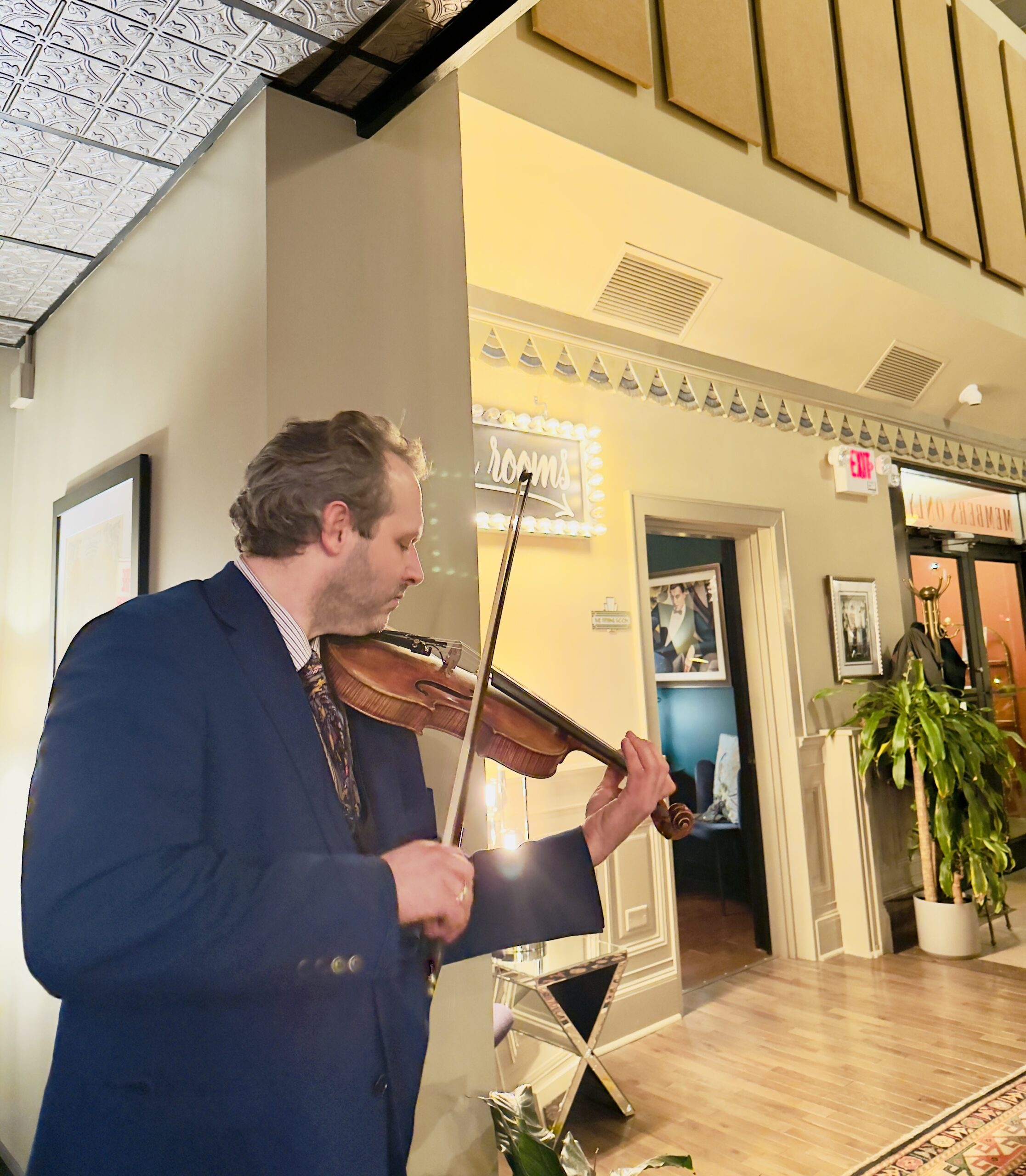 Man playing violin indoors near a plant and framed pictures.